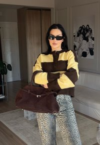 Suéter de malha às riscas castanho e amarelo, calças cinza com padrão de leopardo e uma mala de camurça castanho-escura com um detalhe em prata. Óculos de sol modernos usados.