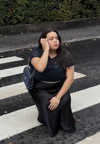 Zwarte satijnen rok, blouse met kant aan de bovenkant, en een gewatteerde marineblauwe handtas. Achtergrond met een donkere stoep en een zebrapad. Geen zichtbare patronen.