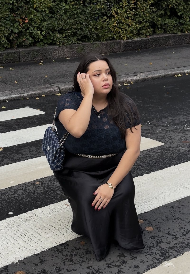 Zwarte satijnen rok, blouse met kant bovenaan, en een gewatteerde marineblauwe handtas. De achtergrond toont een donkere stoep en een zebrapad. Geen zichtbare patronen.