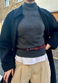 Grå høyhalset genser under hvit skjorte, strammet inn med et rødt skinnbelte, kombinert med beige vidbukser og en svart kåpe.