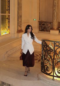 Witte op maat gemaakte blazer over een diep bordeaux satijnen rok, gecombineerd met een kleine bordeaux handtas, op een marmeren trap met sierlijke leuningen.