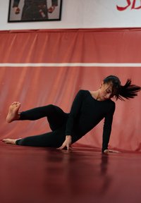 Haut de sport noir à manches longues et leggings portés par une personne sur un tapis rouge, effectuant un exercice avec une jambe levée et pieds nus.