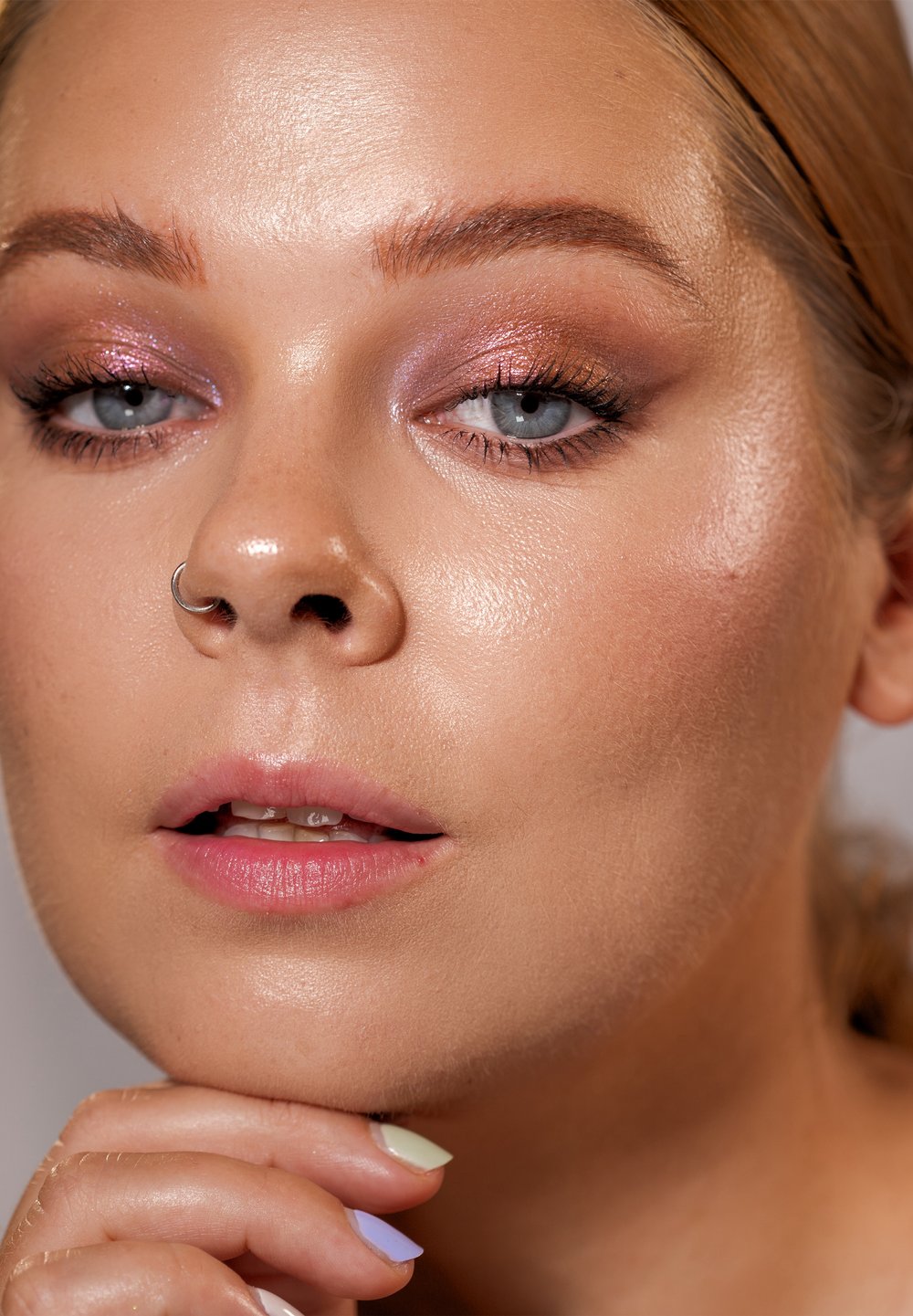 Nærbilde av et ansikt med glatt, strålende hud; blå øyne; lys rosa øyenskygge; nesering; myke rosa lepper; hånd med pastellfargede negler.
