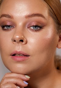 Nærbilde av et ansikt med glatt, strålende hud; blå øyne; lys rosa øyenskygge; nesering; myke rosa lepper; hånd med pastellfargede negler.