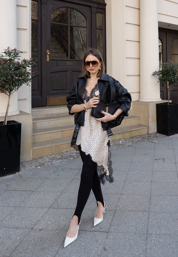 Kvinde iført solbriller, sort læderjakke, hvid prikket kjole med blonder, sorte leggings og hvide hæle, der holder en sort taske udenfor en bygning.
