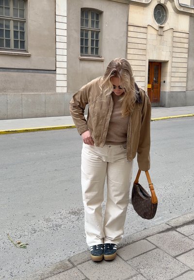 Chaqueta bomber marrón claro, suéter beige, pantalones anchos blancos y zapatillas azul marino con suelas marrón claro. Lleva un bolso oscuro con una correa marrón claro.