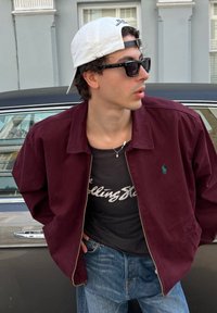 Giovane uomo con berretto bianco indossato all'indietro, occhiali da sole neri, giacca bordeaux, t-shirt dei Rolling Stones e jeans, appoggiato a un'auto.