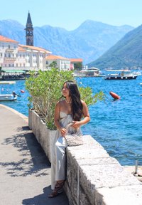 Hellgraues Kleid mit einer Knotendetail, hält eine geflochtene Tasche, neben einer Steinmauer am blauen Wasser, mit Grünflächen und Bergen im Hintergrund.