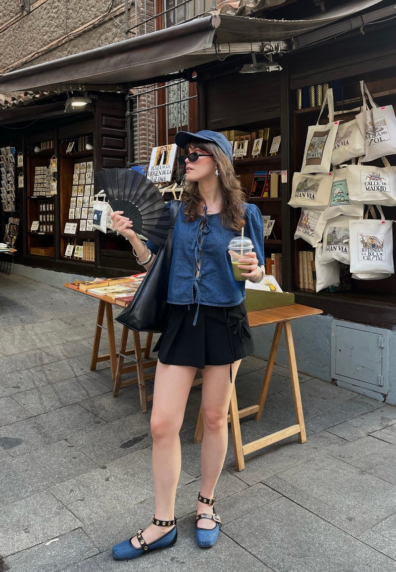 Denimbluse mitBinder, schwarzer Rock, schwarze Fächer, dunkle Sonnenbrille und blaue Schuhe mit Riemen. Schwarze Tragetasche und ein Getränk in der Hand.