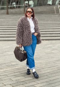 Manteau en fausse fourrure marron foncé, chemise blanche à boutons, jean en denim bleu clair, mocassins noirs avec chaussettes, et sac à main marron foncé.