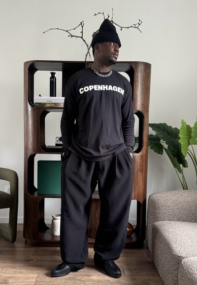 Hombre que lleva una camiseta negra con la palabra "COPENHAGEN", gorro negro y pantalones holgados, de pie en el interior cerca de una estantería de madera y una planta verde.