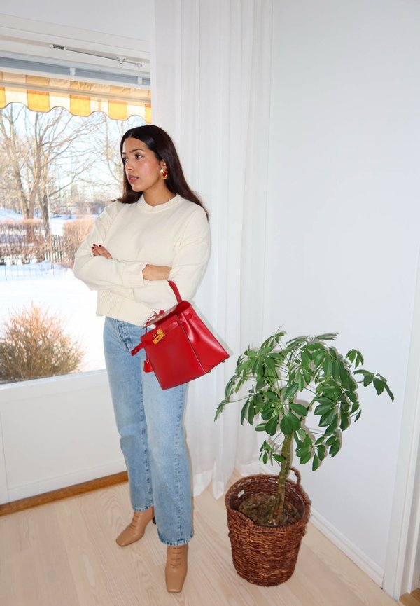 Femme aux longs cheveux foncés portant un pull blanc, un jean bleu, des chaussures beige, tenant un sac à main rouge, debout près d'une plante en pot près d'une fenêtre avec un paysage enneigé à l'extérieur.