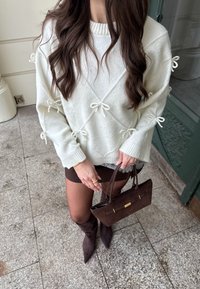Femme portant un pull blanc avec des nœuds, une jupe noire, des collants noirs et des bottes noires à bouts pointus, tenant un sac à main marron foncé.