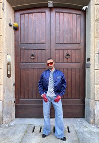 Giacca bomber in satin blu navy, camicia bianca a motivi, cravatta grigia, jeans a zampa d'elefante blu, guanti rossi, occhiali da sole rossi oversized, in piedi accanto a porte in legno.