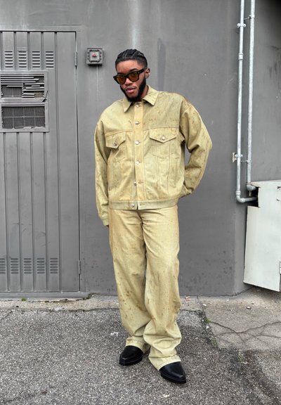 Chaqueta de mezclilla de color amarillo claro-marrón con dos bolsillos frontales y pantalones a juego. Unos zapatos negros de corte slim completan el conjunto frente a una pared gris.