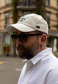 Cappellino da baseball beige con la scritta "La Scelta Intelligente" in nero e il logo New Balance, indossato con occhiali da sole neri e una camicia bianca con colletto.