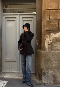 Čierny elegantný blejzer, svetlo-modré rozšírené džínsy, štruktúrovaný čierny klobúk, drží tmavú kabelku, v pozadí so sivými dverami a kamennou stenou.
