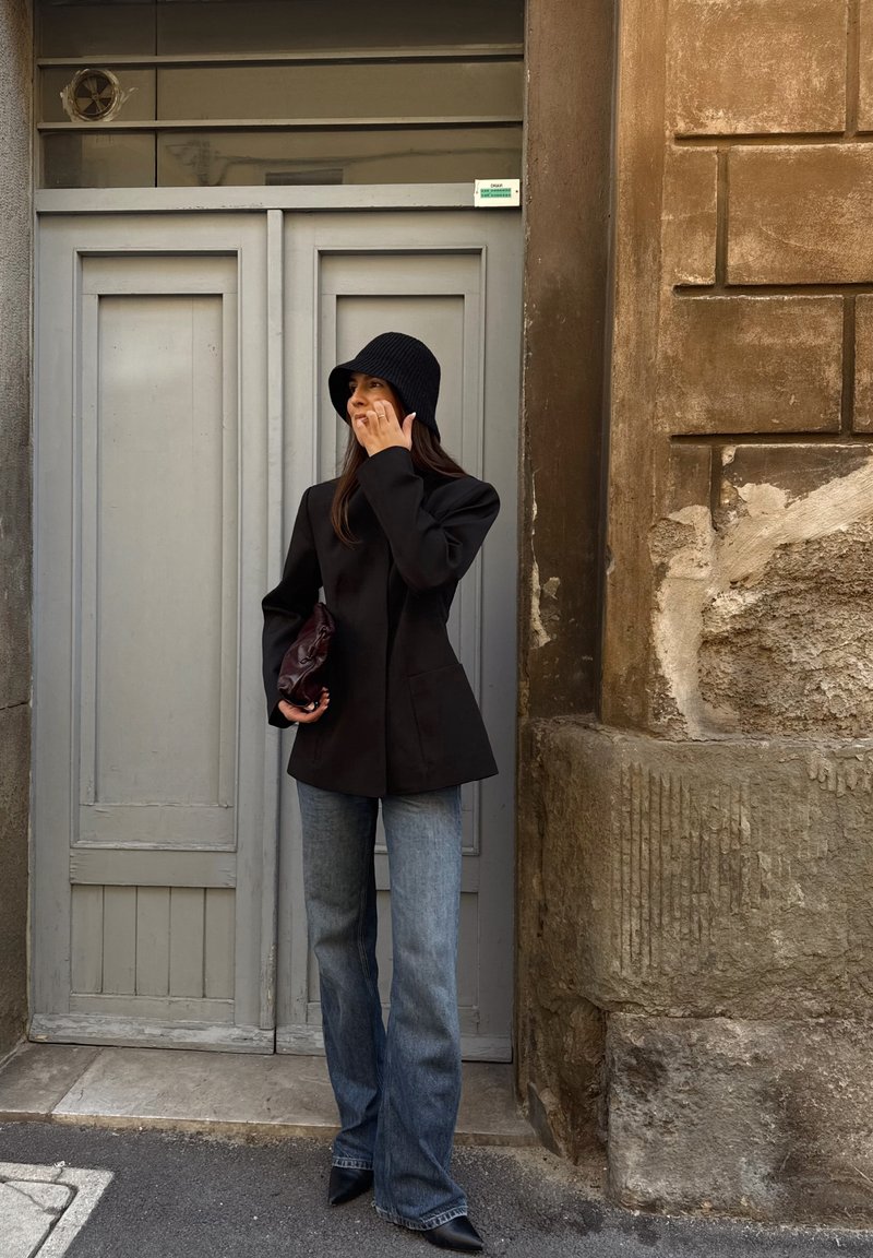 Black tailored blazer, light blue flared jeans, textured black bucket hat, holding a dark handbag, with a grey door and stone wall in the background.