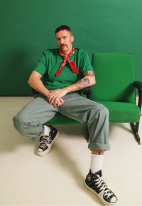 T-shirt de algodão verde, bandana vermelha, calças cinzentas, sapatilhas pretas. Sentado em cadeiras verdes, fundo neutro, tatuagens visíveis nos braços.