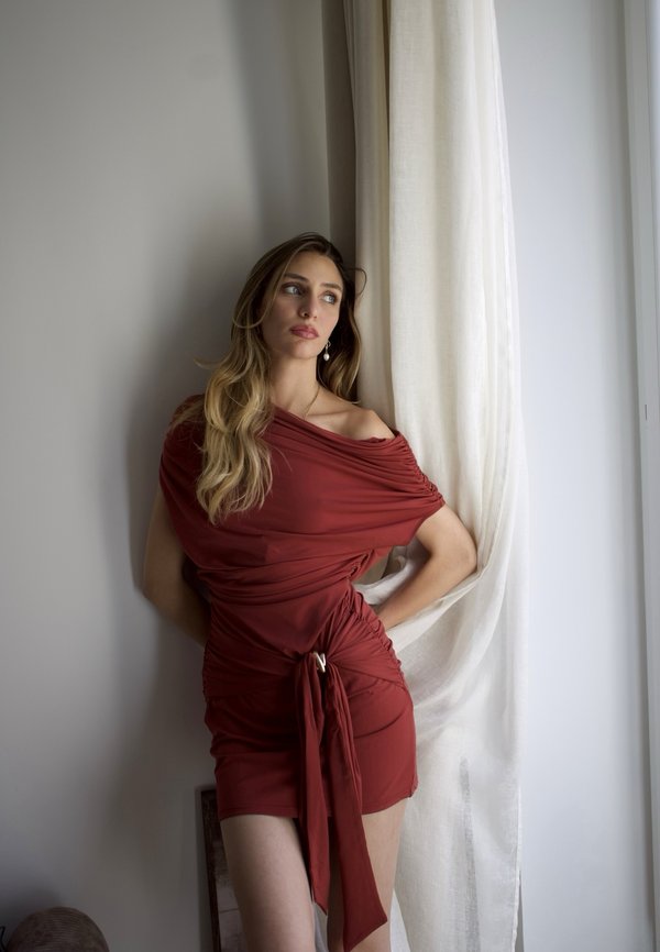 Femeie într-o rochie mulată de culoare ruginiu fără umeri, stă lângă o perdea diafană, uitându-se gânditoare în lateral, cu mâinile pe șolduri.