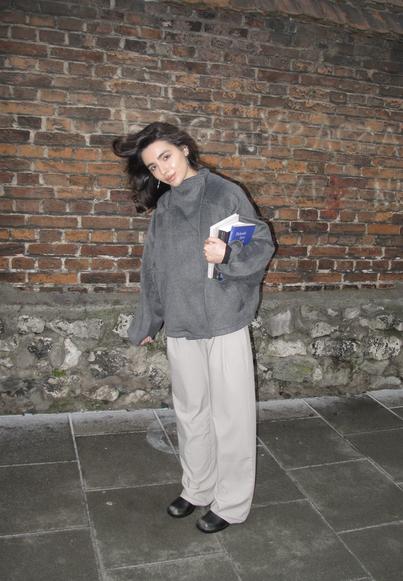 Νεαρή γυναίκα με γκρι φαρδιά μπουφάν και μπεζ παντελόνι κρατώντας βιβλία στέκεται σε λιθόστρωτο δάπεδο μπροστά σε τοίχο από τούβλα τη νύχτα.