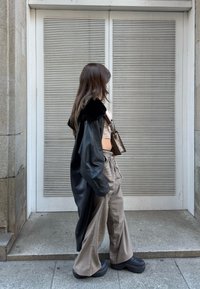 Jeune femme portant un long manteau en cuir noir, un pantalon large à carreaux, des chaussures à plateforme et un petit sac à bandoulière marron, marchant devant une porte métallique fermée.