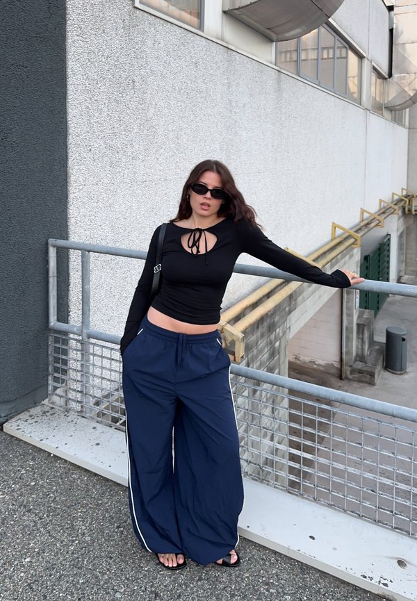 Joven mujer con top negro y pantalones anchos azul marino, gafas de sol, apoyada en una barandilla de metal fuera, junto a una pared de hormigón.