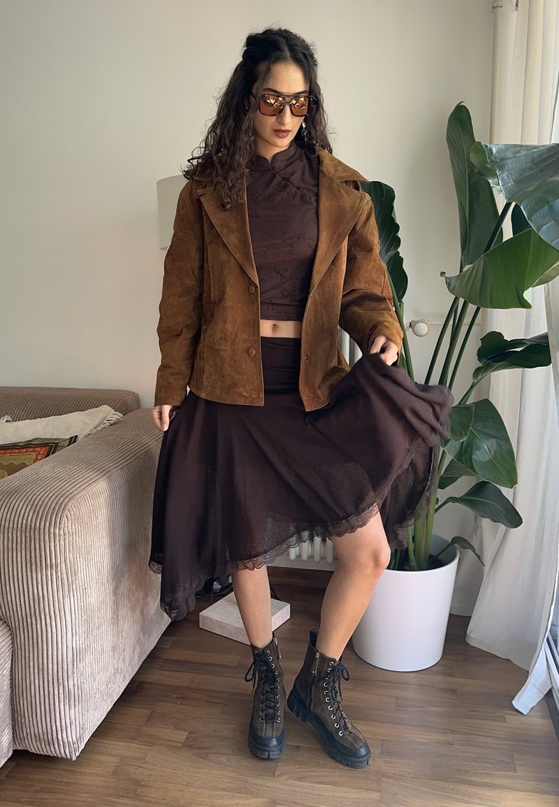 Donna che indossa una giacca in suede marrone, un crop top marrone scuro, una gonna asimmetrica, stivali con lacci e occhiali da sole all'interno vicino a una pianta e un divano.