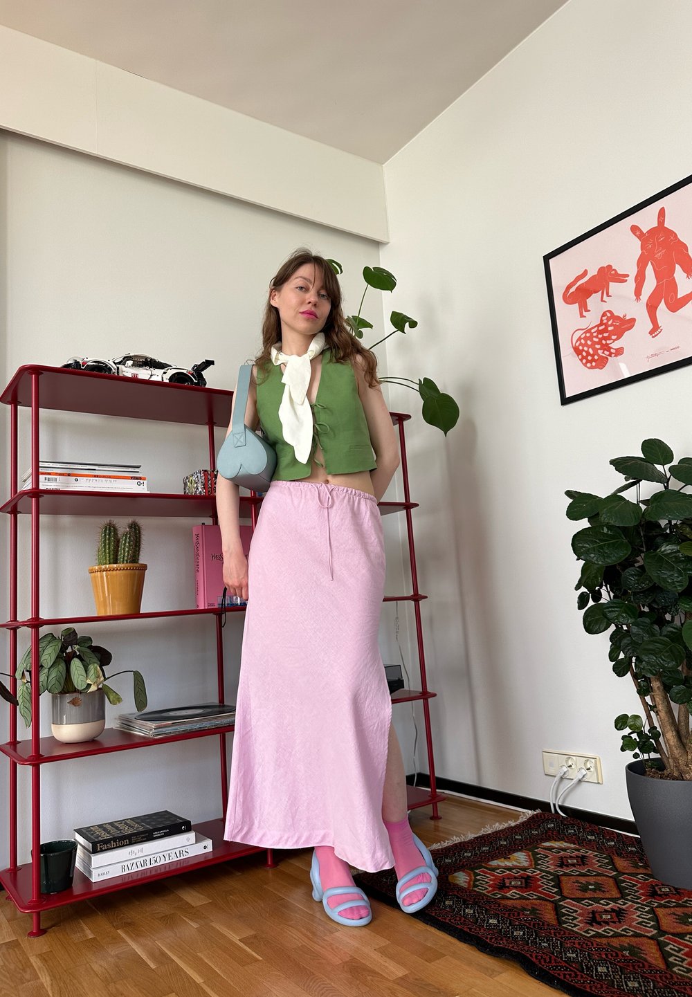 Top cropped verde, saia maxi rosa, bolsa azul e sandálias de plataforma azuis. Prateleiras com livros e plantas em um ambiente interior moderno.