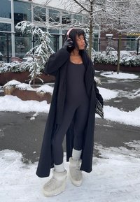Long manteau noir sur un justaucorps noir, chaussettes en tricot gris et bottes de neige blanches. Porte des écouteurs roses. Fond enneigé avec des arbres.