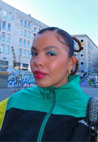 Femme aux cheveux tressés portant un eyeliner bleu, un rouge à lèvres rose, des boucles d'oreilles créoles dorées, une veste Adidas verte et noire, debout dans un quartier urbain avec des graffitis.