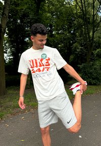 T-shirt Nike blanche avec des lettres rouges et short gris. La personne s'étire en portant des chaussures de sport blanches et rouges dans un parc vert.