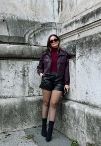 Κοντό μπουφάν από μπορντό δέρμα πάνω από ένα κόκκινο πουλόβερ με ψηλό λαιμό, συνδυασμένο με μαύρες ψεύτικες δερμάτινες βερμούδες και μαύρες κάλτσες μέχρι τον αστράγαλο, σε φόντο μια πέτρινη τοιχοποιία.