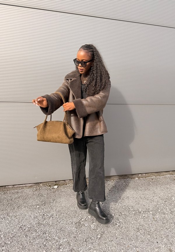 Sieviete, tērpusies brūnā kažokādas virsjakā, melnās biksēs un zābakos, rokās turot brūnu velūra somu pie pelēkas gofrētas sienas.