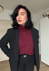 Blazer noir sur mesure porté sur un pull à col roulé bordeaux, associé à un pantalon noir. Boucle de ceinture dorée notable avec des initiales.