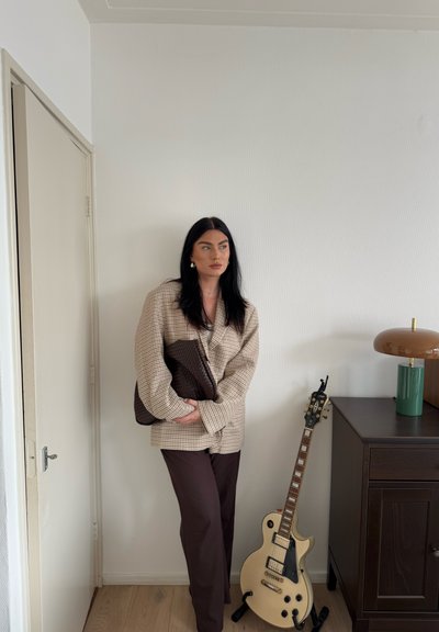 Blazer beige de gran tamaño a cuadros, pantalones marrón oscuro y un bolso marrón texturizado. El fondo incluye una guitarra y una lámpara sobre un armario de madera.