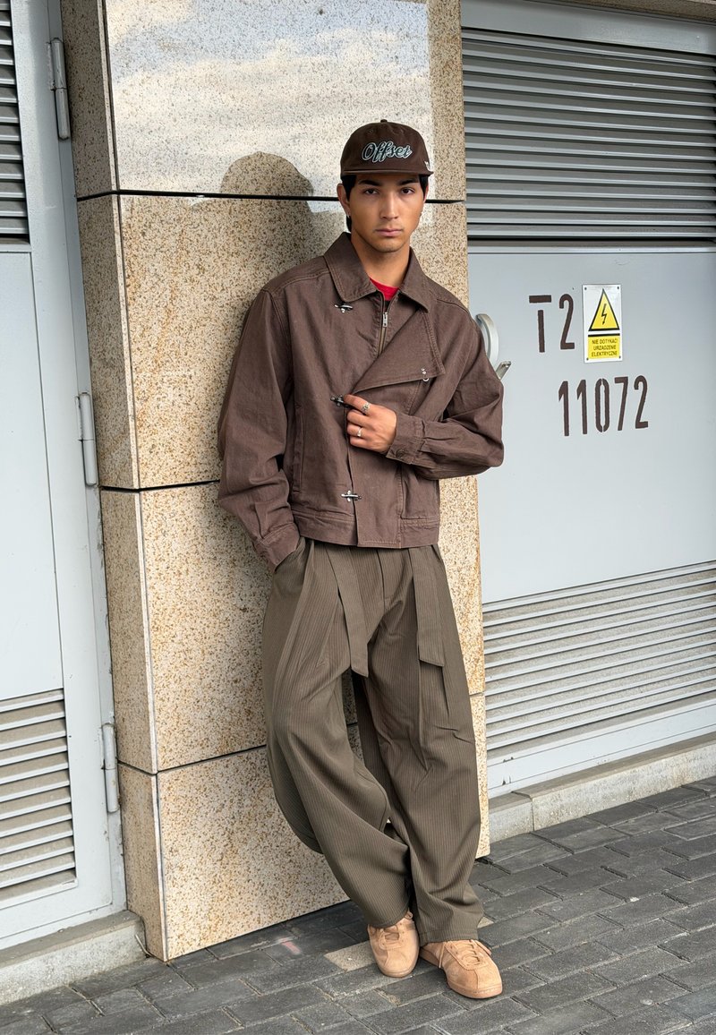 Jeune homme portant une veste marron, une casquette et un pantalon marron ample, appuyé contre un mur en pierre près d'un coffret électrique à l'extérieur.