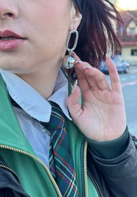 Orecchini a cerchio oversize in argento con un charm sferico. Il primo piano mostra una superficie liscia e riflettente contro una giacca verde e una cravatta a quadri.