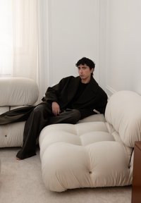 Sofa krem boje, sa jastučićima i zaobljenim rubovima. Unutarnji prostor ima svjetlost iz prozora i neutralne tonove. Osoba u tamnoj odjeći sjedi.