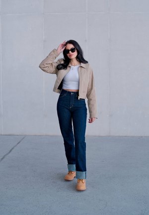 Mujer con cabello largo y oscuro que lleva gafas de sol, chaqueta beige, top corto blanco, vaqueros oscuros con puños enrollados y botas color canela, de pie contra una pared lisa.