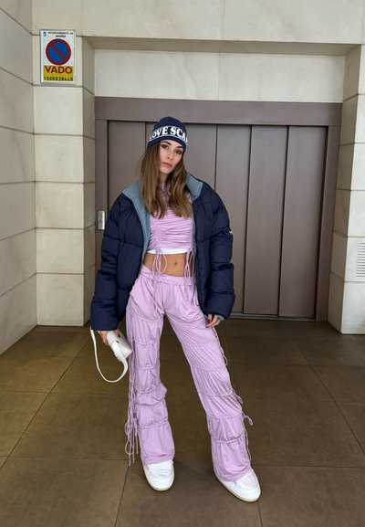 Chaqueta de plumón en azul oscuro con forro gris, top corto arrugado rosa, pantalones de tiro alto en rosa claro con cordón, y zapatillas blancas gruesas, sosteniendo un bolso blanco.