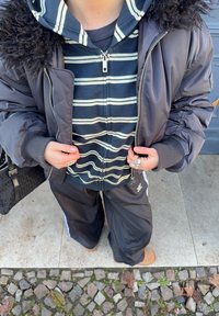 Svart polstret jakke med fuskepelskrage, stripete marineblå og hvit hettegenser under, svarte joggebukser og brune sko. Ring på høyre hånd.