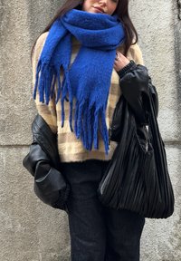 Žena v béžovom pruhovanom svetri, s veľkým modrým šálom, čiernymi nohavicami a čiernou skladanou kabelkou, stojaca pri textúrovanej betónovej stene.