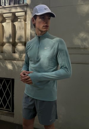 Camisa atlética de cremallera en color teal claro con mangas largas y un patrón texturizado. Combinada con pantalones cortos grises que tienen un pequeño logo de Nike y una gorra blanca.