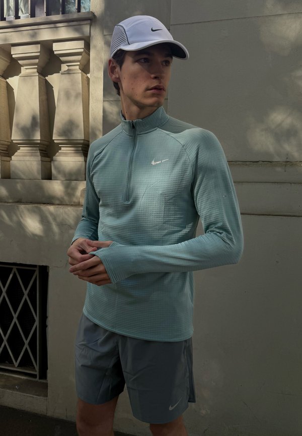Lichtblauw groene zip-up sportshirt met lange mouwen in een gestructureerd patroon. Gecombineerd met grijze shorts met een klein Nike-logo en een witte pet.