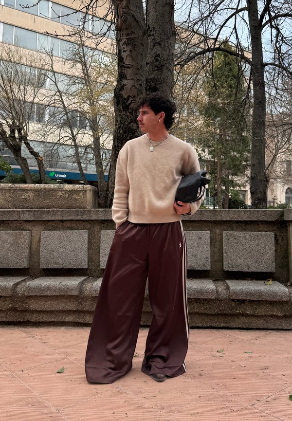 Tânăr cu păr creț purtând pulover bej și pantaloni largi maro, ținând o geantă neagră, stând lângă o bancă din piatră și copaci goi.