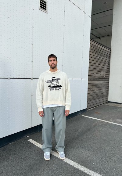 Suéter de punto crema con texto gráfico negro, superpuesto a una camisa blanca; pantalones de pierna ancha en gris claro; zapatillas grises; de pie frente a un fondo texturizado.