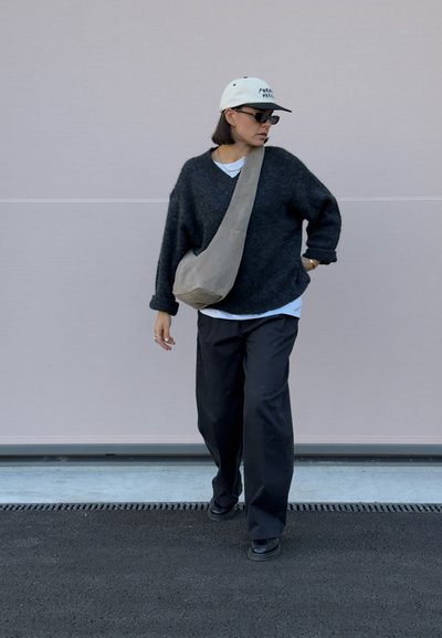 Jersey de lana negro de gran tamaño, camiseta blanca debajo, pantalones negros holgados, bolso cruzado gris, gorra blanca, gafas de sol, de pie sobre el pavimento.