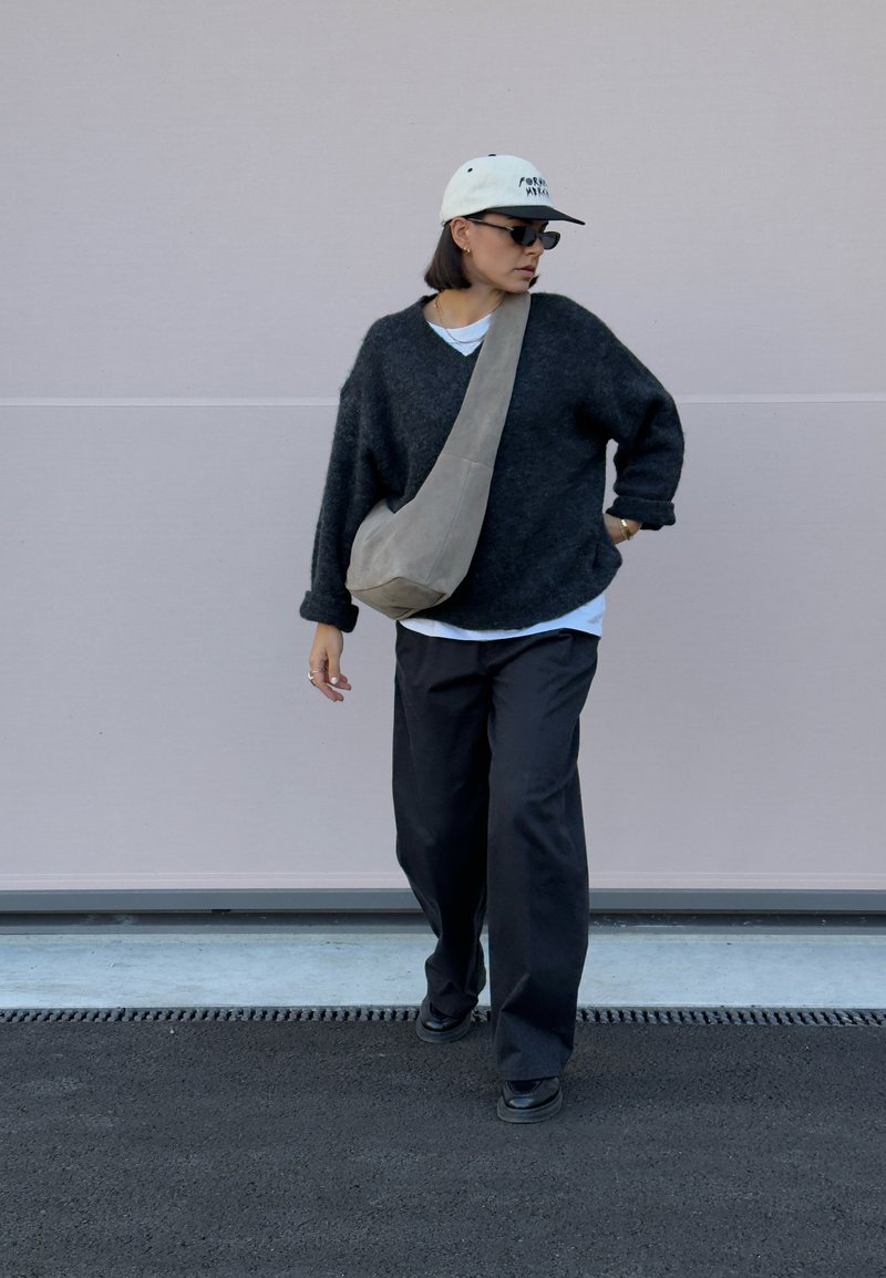 Pulôver de lã oversized preto, t-shirt branca por baixo, calças pretas descontraídas, mala a tiracolo cinza, boné branco, óculos de sol, a pé no pavimento.