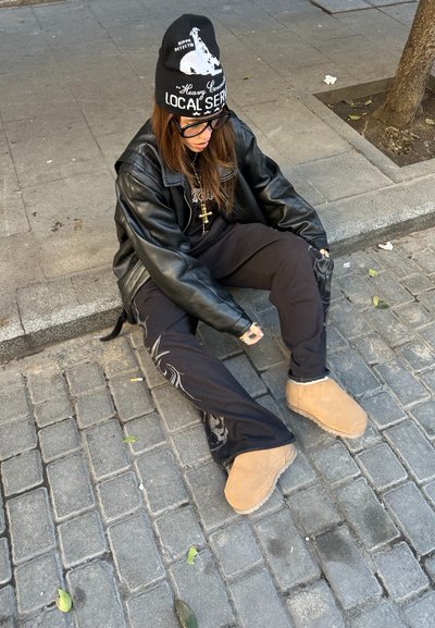 Individual sentado en una acera, llevando una chaqueta de cuero negra, pantalones negros con un patrón plateado y botas de piel de imitación de color marrón claro. Gorra negra y gafas.